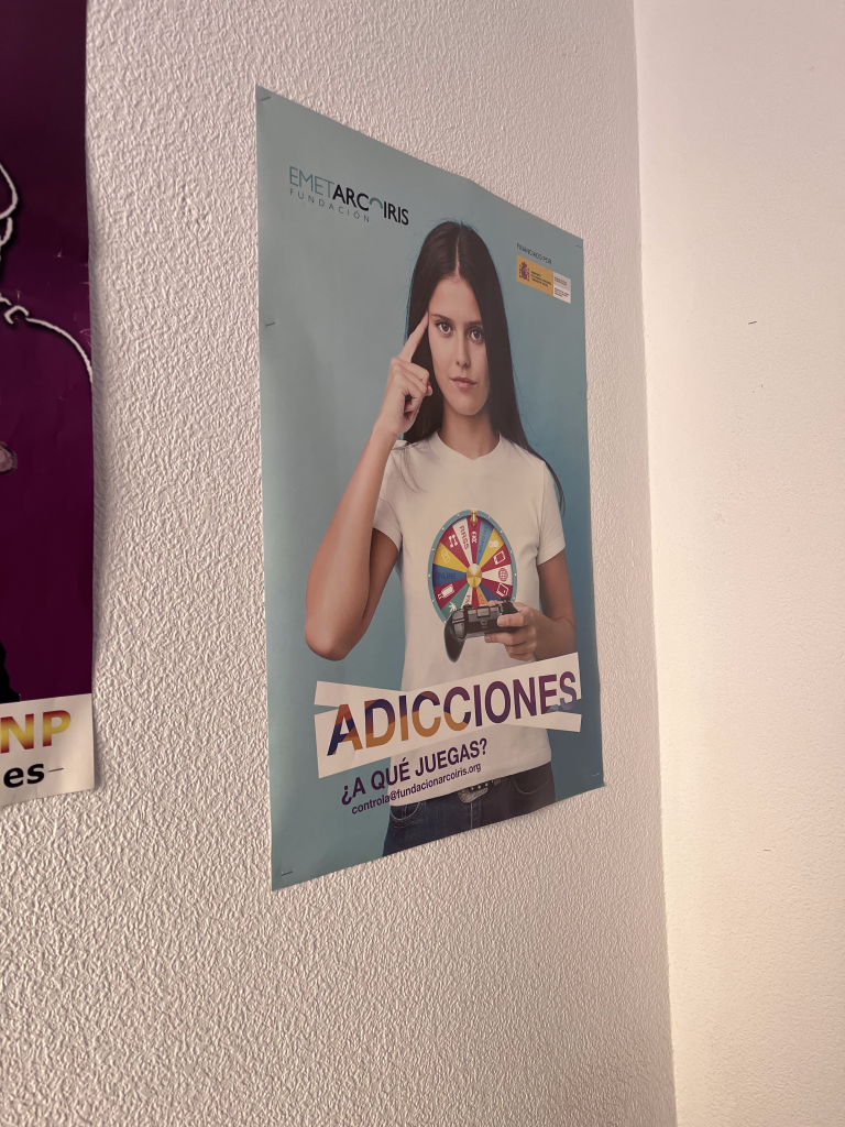 Plakat fundacji EMET Arco Iris przedstawiający młodą kobietę z kontrolerem do gier i napisem „ADICCIONES. ¿A QUÉ JUEGAS?” (Uzależnienia. W co grasz?).