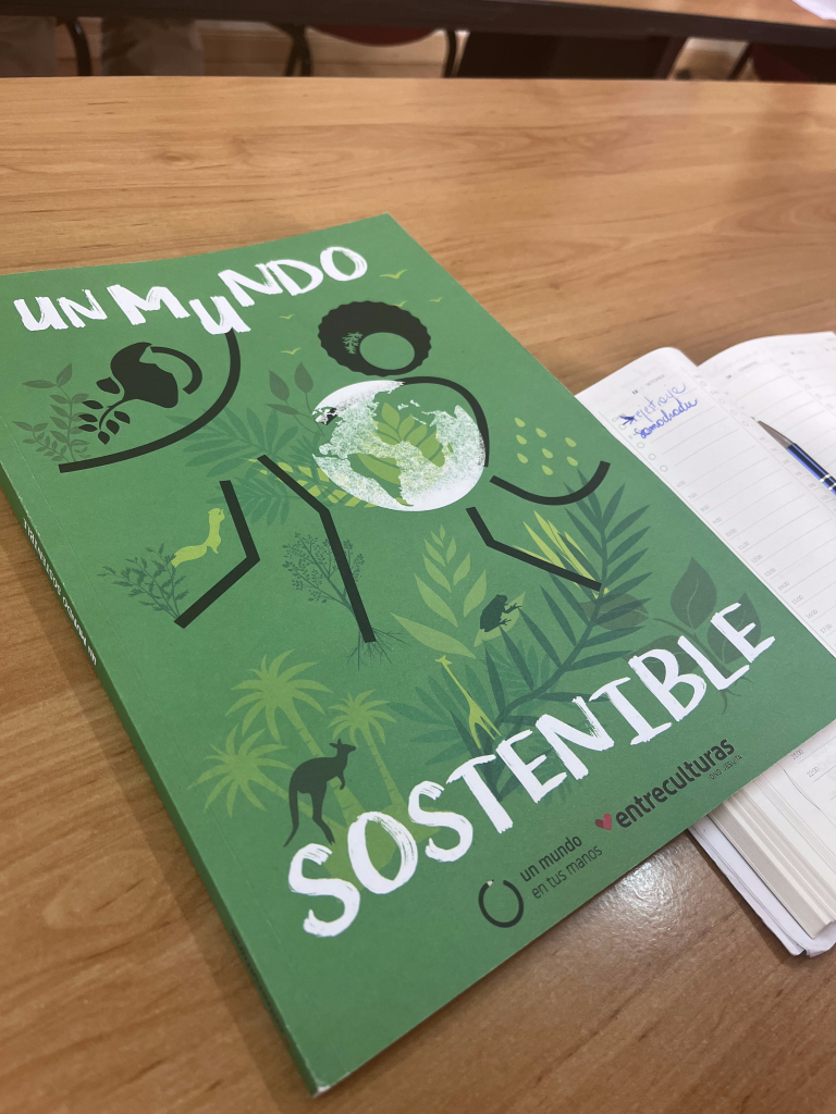 Zielona książka pt. „UN MUNDO SOSTENIBLE” leżąca na drewnianym stole obok otwartego kalendarza. Na okładce ilustracja globu wkomponowana w sylwetkę człowieka otoczonego roślinnością i zwierzętami.